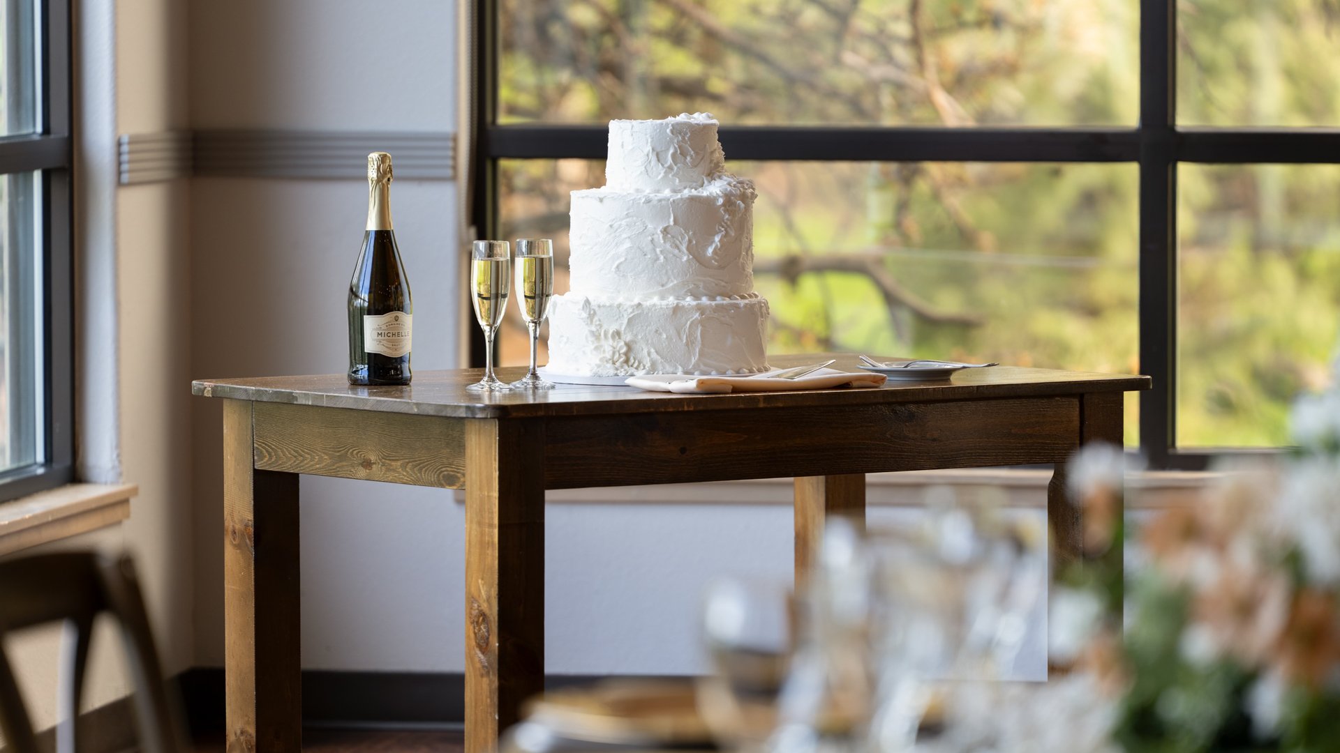 Rustic wooden table displaying a white tiered wedding cake and champagne glasses beside large windows with mountain views at The Pines reception.