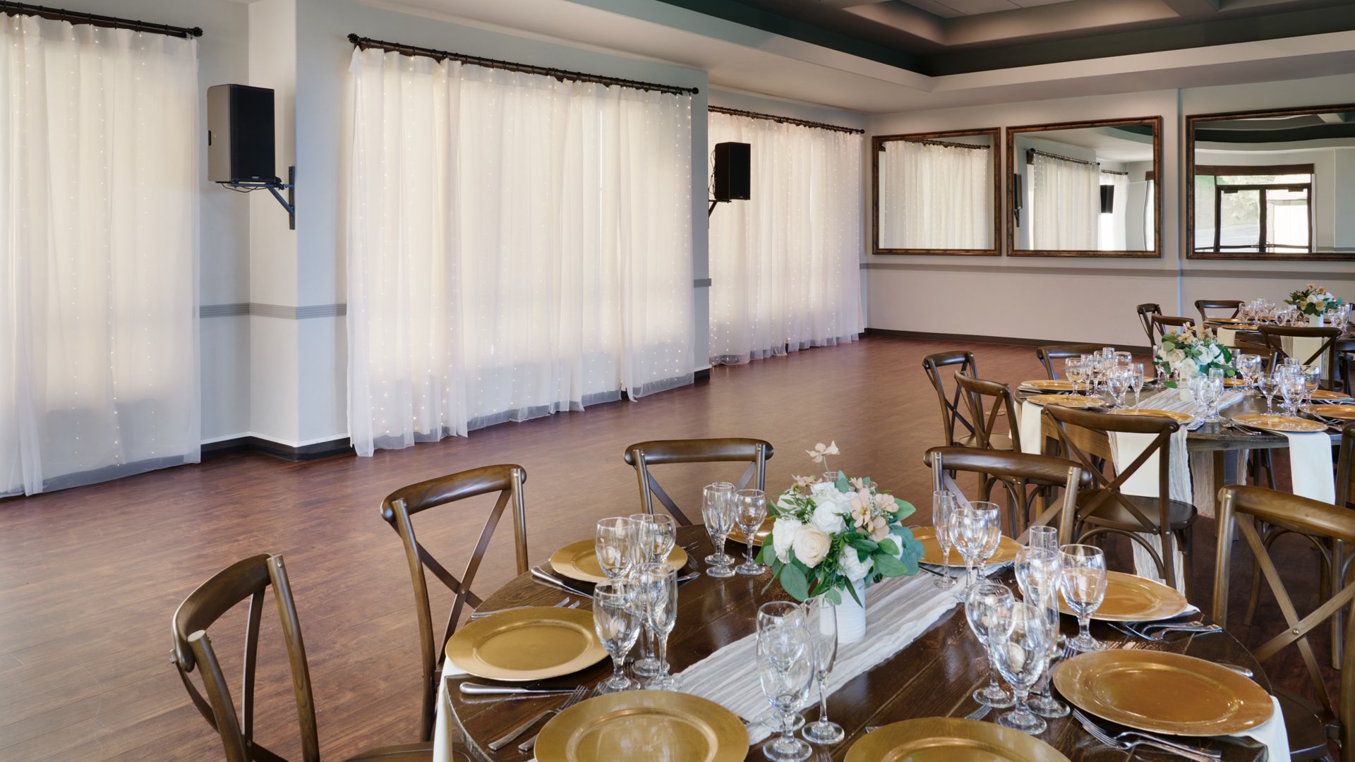 Wedding reception room at The Pines with round tables set with gold chargers, white floral centerpieces, wooden cross-back chairs, and sheer draped curtains.