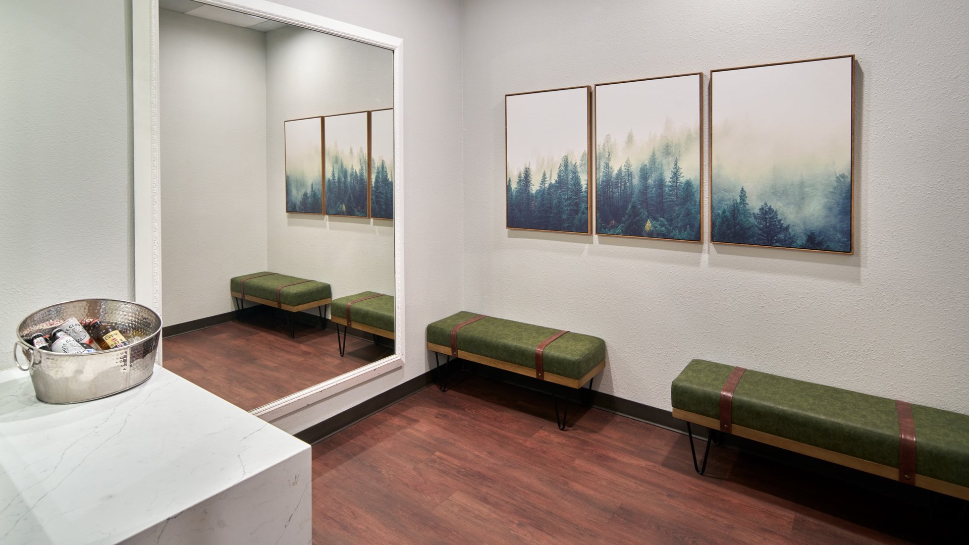 Stylish groom’s lounge featuring forest-themed wall art, green benches, a large mirror, and a silver beverage bucket with drinks at The Pines.