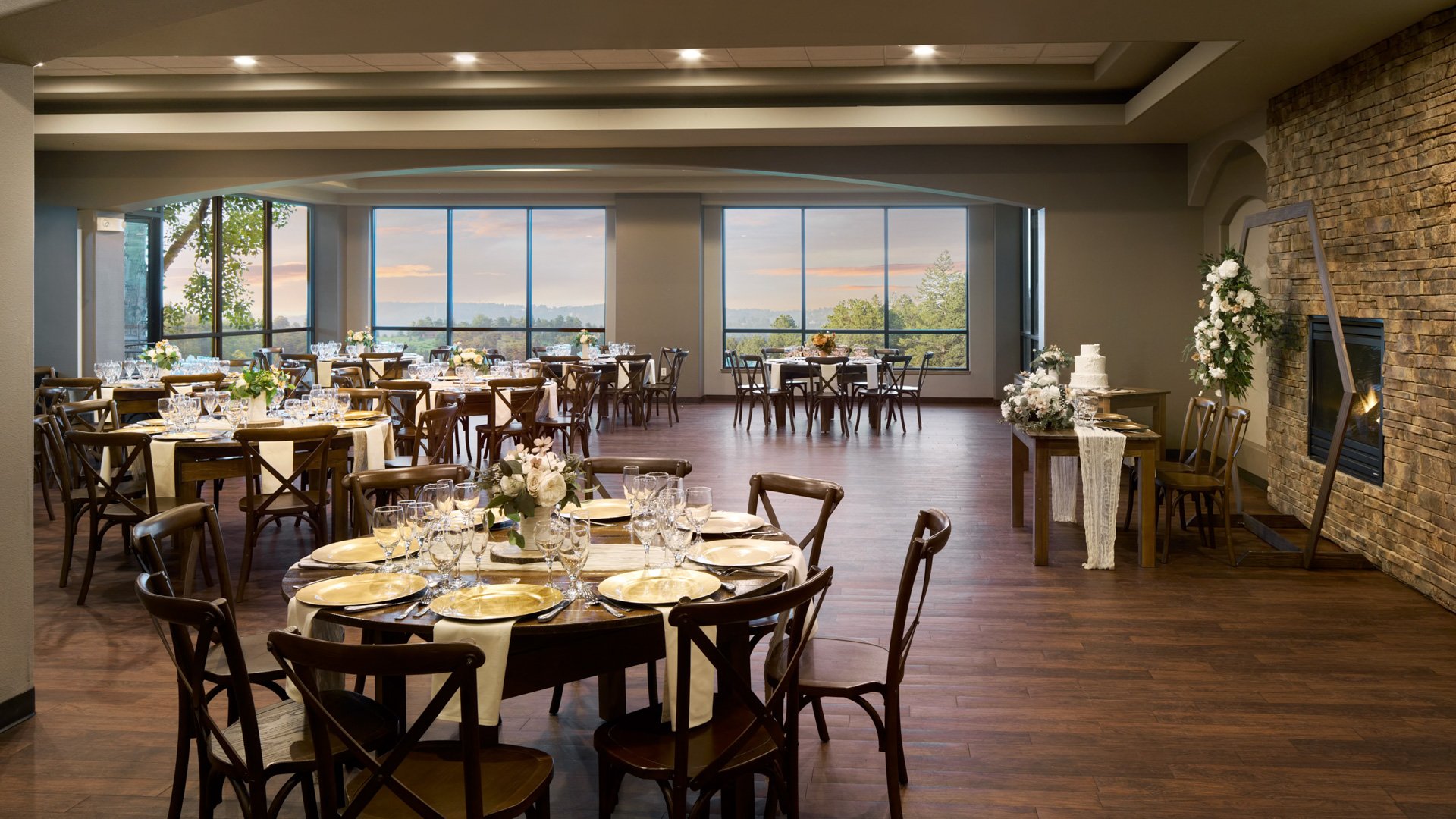 Expansive wedding reception ballroom at The Pines showcasing floor-to-ceiling windows, wooden floors, elegant table settings, and a stone fireplace with floral décor.