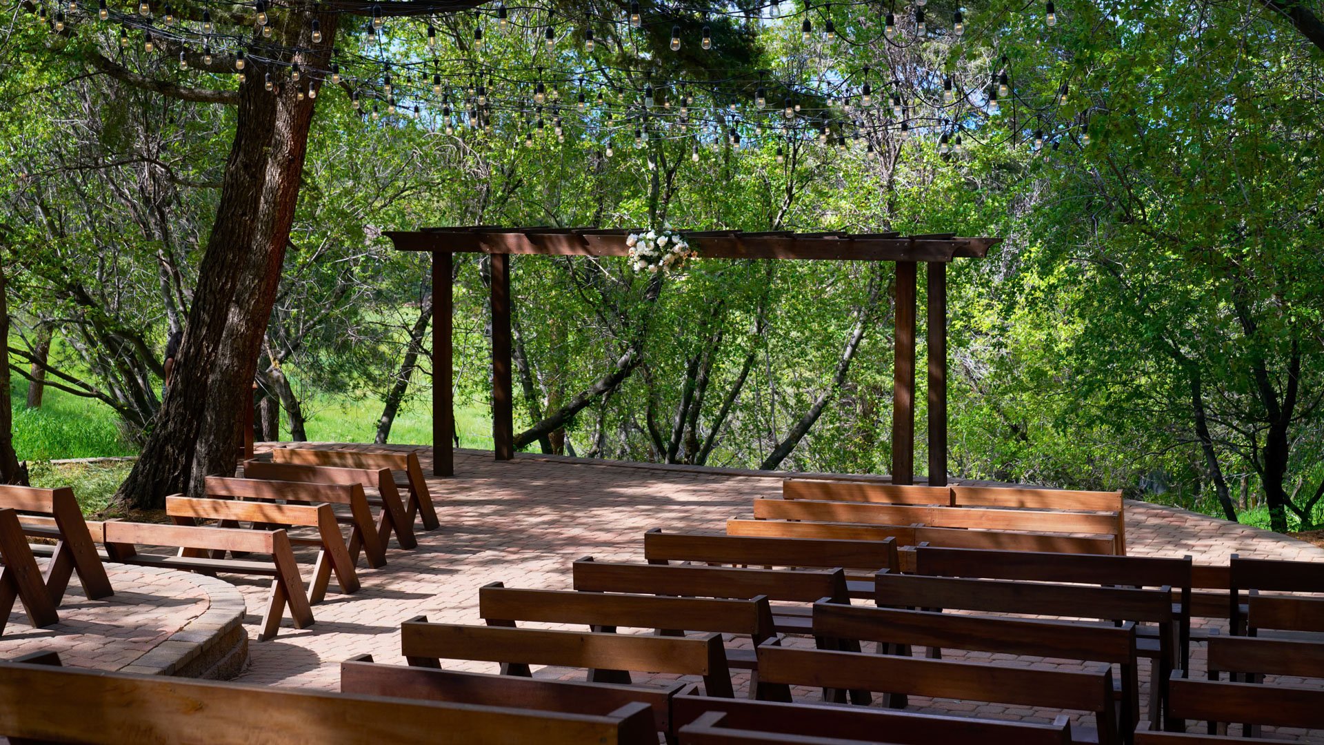 Close-up view of The Pines Aspen Terrace with wooden benches, a floral-accented arch, and string lights under a canopy of green trees.