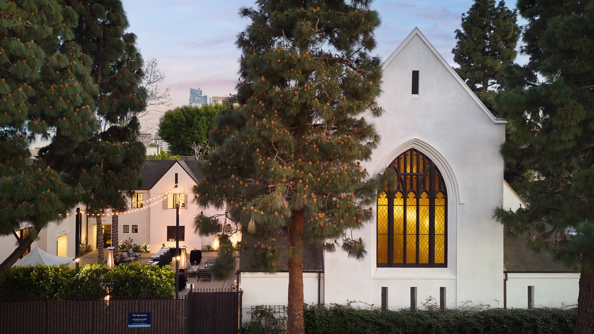 Exterior view of The Sanctuary Cathedral in Long Beach at dusk, featuring illuminated stained glass and courtyard lighting.