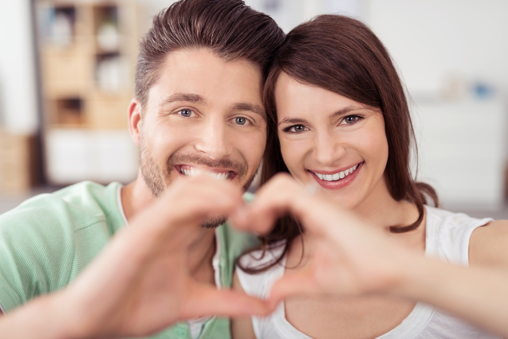 Close up Happy Sweet Young Couple at the Living Room Showing Heart Shape using Hands and Smiling at the Camera.