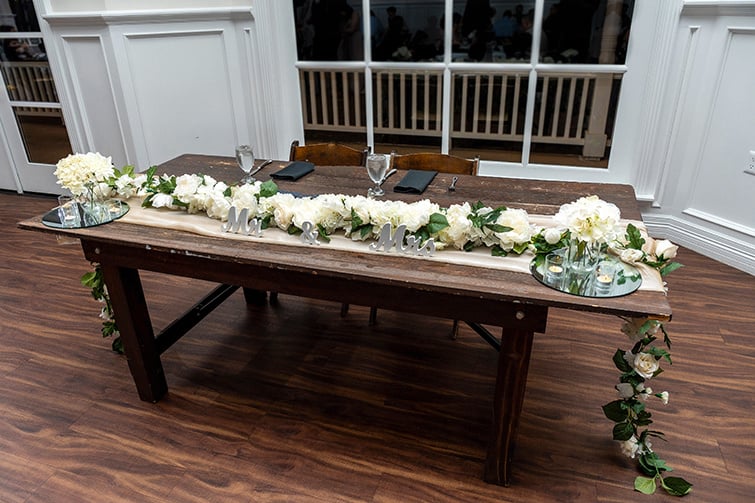 A wedding table setting adorned with a floral garland and elegant centerpieces featuring white flowers and greenery.