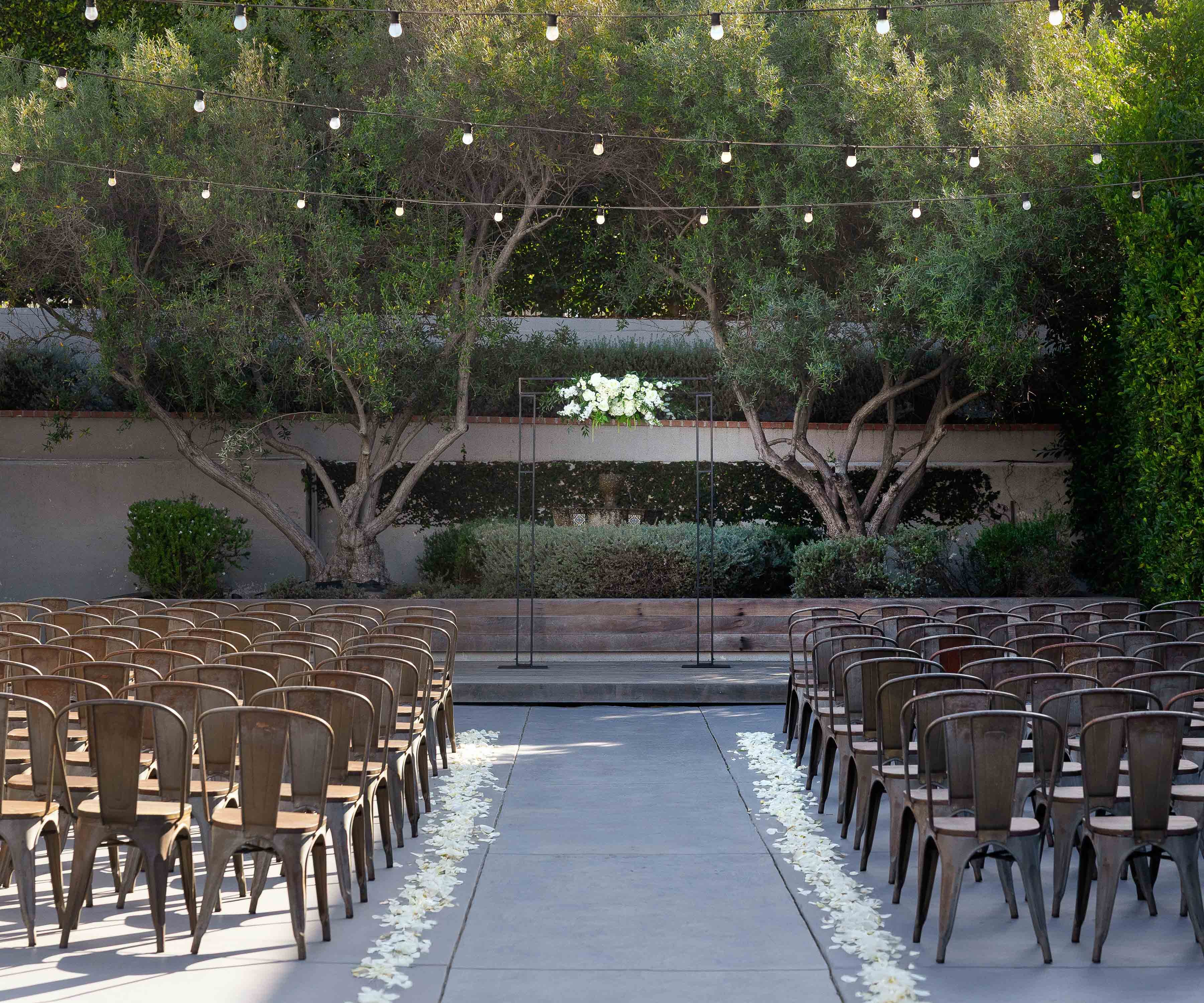 Outdoor wedding ceremony space at The Harper wedding venue in Costa Mesa, California. Metal bistro chairs are arranged on either side of a central aisle scattered with white flower petals. At the end of the aisle stands a minimalist metal arch adorned with white floral arrangements.