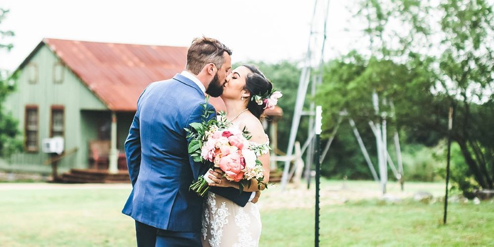 Simple Outdoor Texas Wedding: A newlywed couple kissing outdoors, surrounded by trees and a barn close-by.
