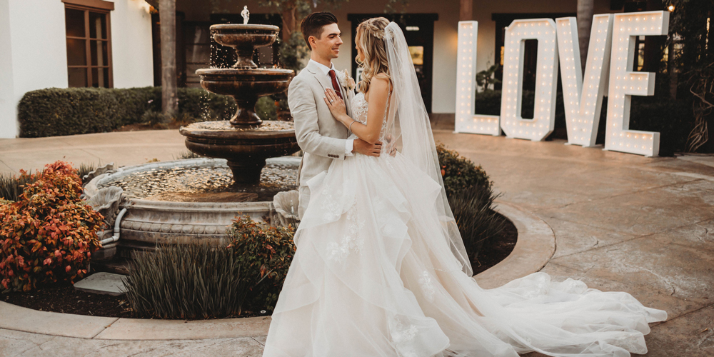 Maria & Hayden standing in the courtyard at Fallbrook Estate next to a fountain and the marquee love letters