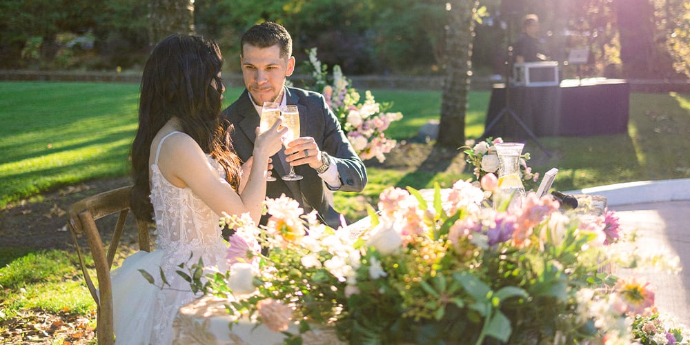 A bride and groom toast with champagne at an outdoor wedding reception table adorned with flowers, set against a sunny, natural backdrop.